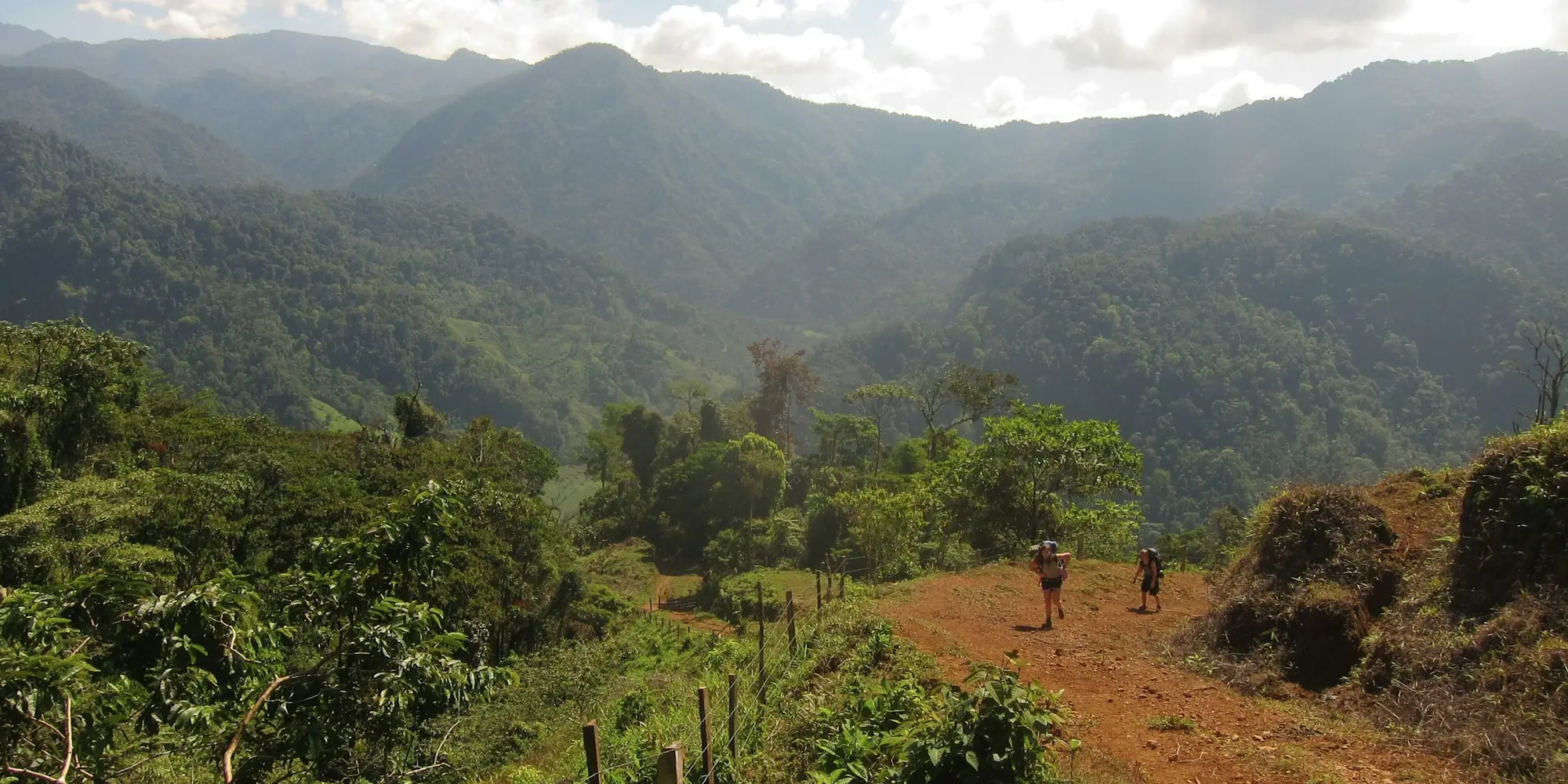 Two people walking a red-earth track along a fence line into forested hills in Costa Rica.